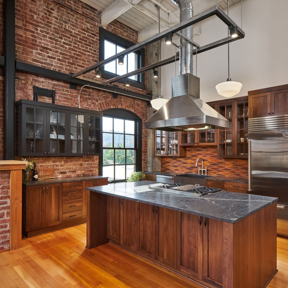 Mid-sized industrial l-shaped open plan kitchen in Portland with a farmhouse sink, glass-front cabinets, dark wood cabinets, soapstone benchtops, multi-coloured splashback, ceramic splashback, stainless steel appliances, medium hardwood floors, with island, brown floor, black benchtop and exposed beam.