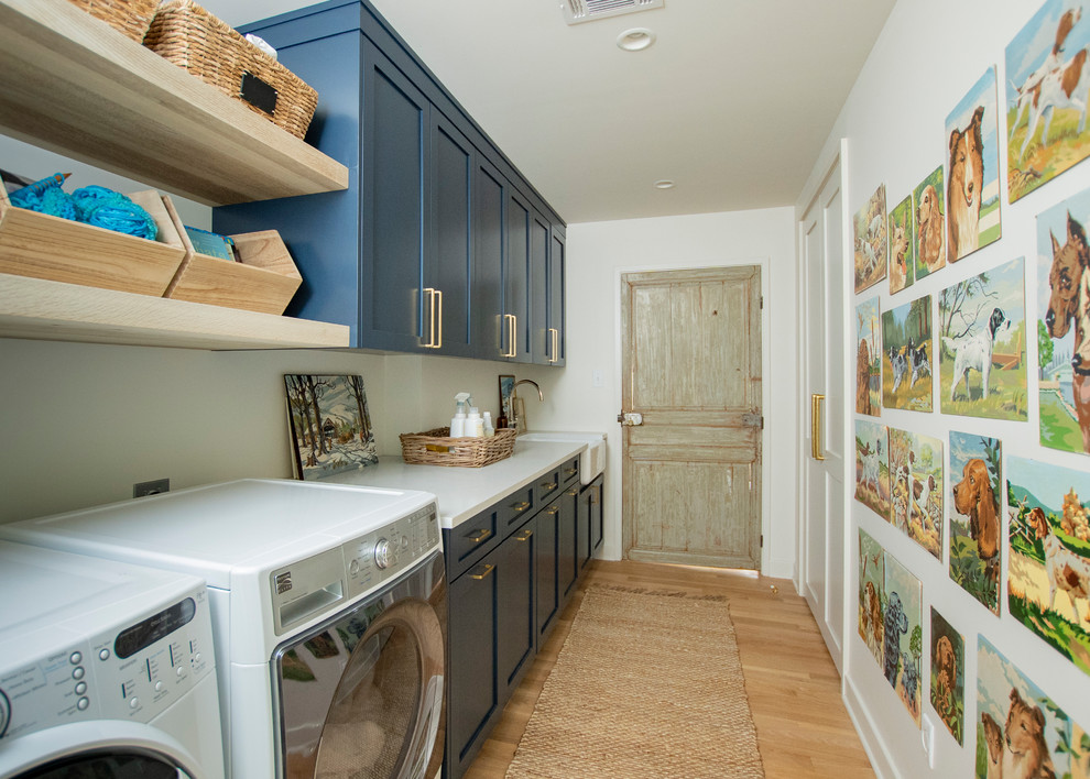 Design ideas for a mid-sized country single-wall dedicated laundry room in Other with a farmhouse sink, shaker cabinets, blue cabinets, quartz benchtops, white walls, a side-by-side washer and dryer, brown floor, white benchtop and medium hardwood floors.
