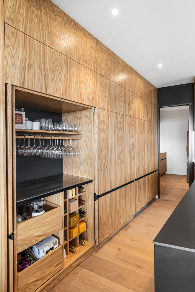 This is an example of a small contemporary wet bar in Auckland with light wood cabinets, stainless steel benchtops, black splashback, porcelain splashback, medium hardwood floors, brown floor, no sink and grey benchtop.