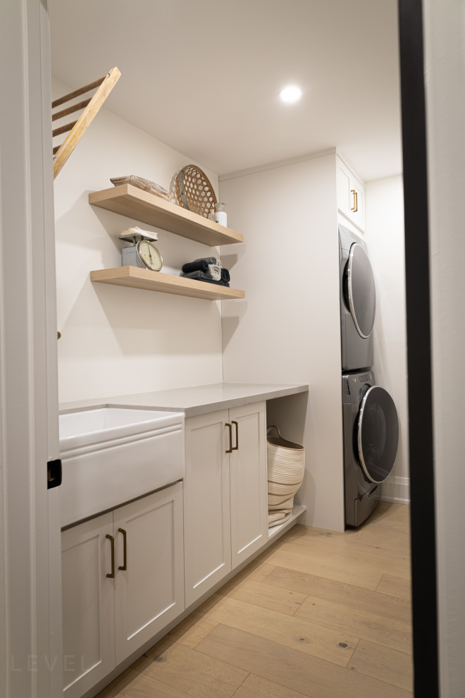 Inspiration for a mid-sized contemporary single-wall dedicated laundry room with a farmhouse sink, shaker cabinets, white cabinets, laminate benchtops, white walls, light hardwood floors, a stacked washer and dryer, brown floor and white benchtop.