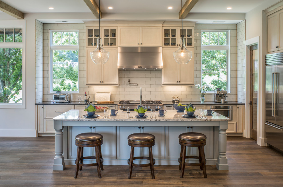This is an example of a large traditional l-shaped eat-in kitchen in Charlotte with a farmhouse sink, flat-panel cabinets, beige cabinets, granite benchtops, white splashback, ceramic splashback, stainless steel appliances, dark hardwood floors, with island, brown floor, black benchtop and exposed beam.