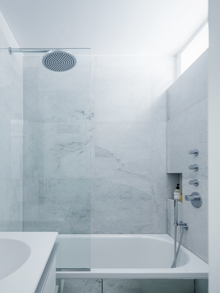 Small modern bathroom in London with flat-panel cabinets, white cabinets, a drop-in tub, an alcove shower, a wall-mount toilet, gray tile, stone tile, white walls, marble floors and a drop-in sink.