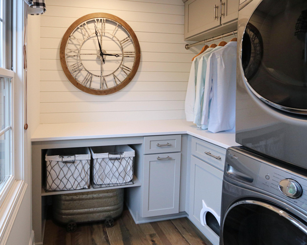 Photo of a small transitional u-shaped dedicated laundry room with recessed-panel cabinets, grey cabinets, quartz benchtops, white splashback, shiplap splashback, grey walls, a stacked washer and dryer and white benchtop.
