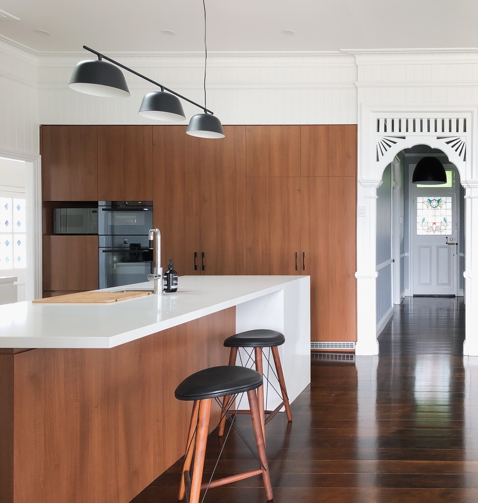 This is an example of a contemporary l-shaped kitchen in Brisbane with flat-panel cabinets, medium wood cabinets, black appliances, medium hardwood floors, with island, brown floor and white benchtop.