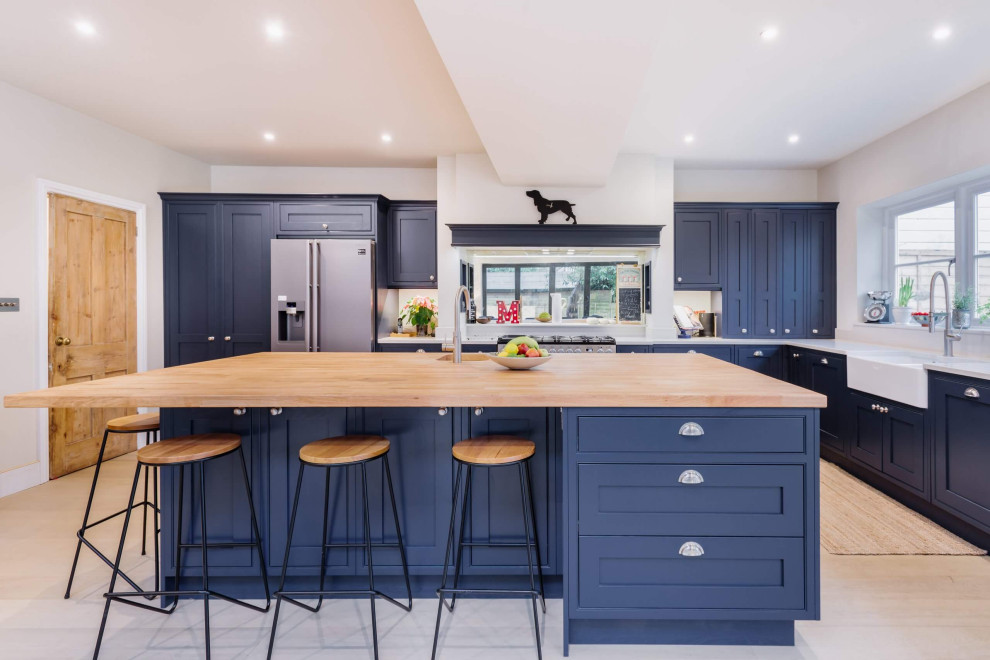 This is an example of a mid-sized transitional u-shaped eat-in kitchen in Sussex with a farmhouse sink, blue cabinets, wood benchtops, stainless steel appliances, with island, brown floor, brown benchtop, shaker cabinets, multi-coloured splashback, mirror splashback and light hardwood floors.