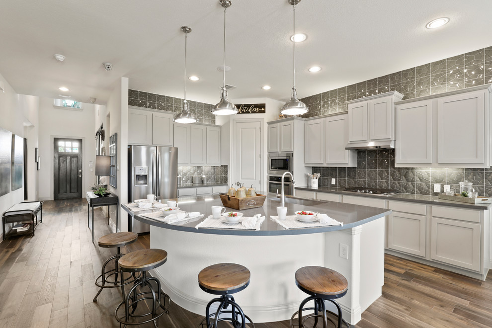 Photo of a large contemporary l-shaped eat-in kitchen in Dallas with a drop-in sink, shaker cabinets, grey cabinets, solid surface benchtops, grey splashback, glass sheet splashback, stainless steel appliances, dark hardwood floors, with island, brown floor and grey benchtop.