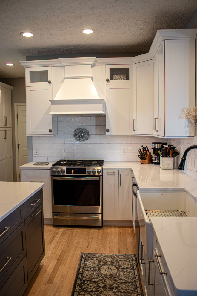 This is an example of a medium sized classic galley kitchen pantry in Cleveland with a belfast sink, shaker cabinets, white cabinets, engineered stone countertops, white splashback, ceramic splashback, stainless steel appliances, light hardwood flooring, an island, brown floors and white worktops.
