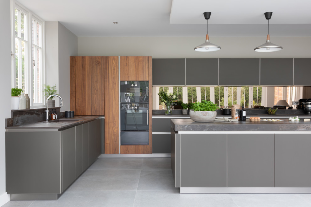 Large contemporary open plan kitchen in Hampshire with an integrated sink, glass-front cabinets, grey cabinets, quartzite benchtops, metallic splashback, mirror splashback, black appliances, with island and grey benchtop.
