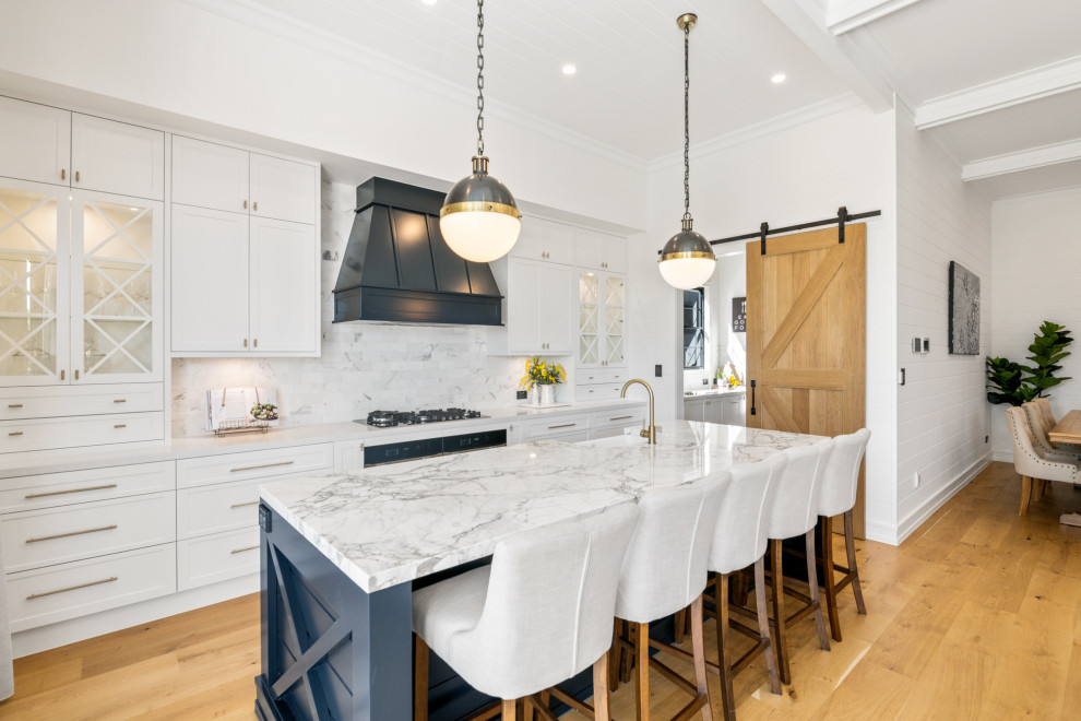 This is an example of a country galley kitchen in Brisbane with a farmhouse sink, shaker cabinets, white cabinets, white splashback, stainless steel appliances, medium hardwood floors, with island, brown floor and white benchtop.
