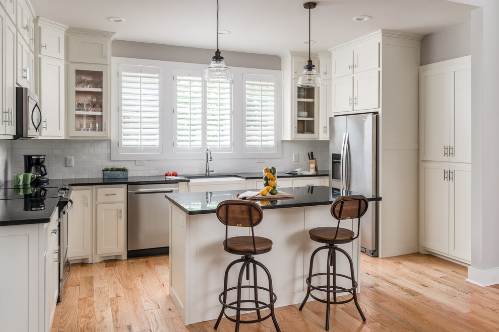 Inspiration for a small transitional l-shaped open plan kitchen in Nashville with a farmhouse sink, shaker cabinets, white cabinets, white splashback, subway tile splashback, stainless steel appliances, light hardwood floors, with island and black benchtop.