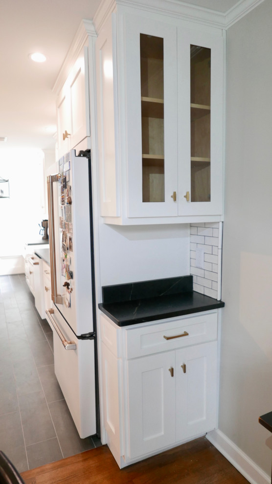 Example of a mid-sized transitional galley porcelain tile and black floor eat-in kitchen design with a farmhouse sink, shaker cabinets, white cabinets, soapstone countertops, white backsplash, subway tile backsplash, white appliances, an island and black countertops