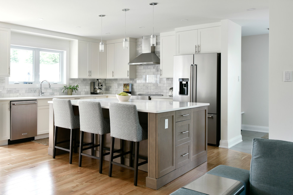 Mid-sized transitional l-shaped open plan kitchen in Ottawa with an undermount sink, shaker cabinets, white cabinets, quartz benchtops, grey splashback, marble splashback, stainless steel appliances, light hardwood floors, with island, beige floor and white benchtop.