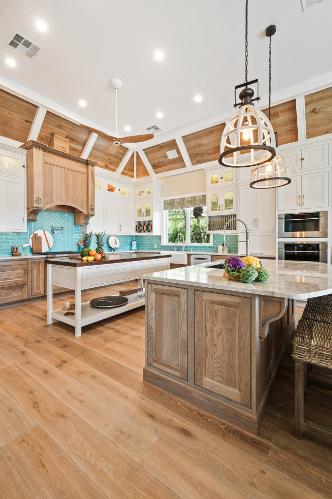 Photo of a large tropical u-shaped open plan kitchen in Other with an undermount sink, beaded inset cabinets, white cabinets, quartzite benchtops, blue splashback, glass tile splashback, stainless steel appliances, light hardwood floors, multiple islands, brown floor, beige benchtop and wood.
