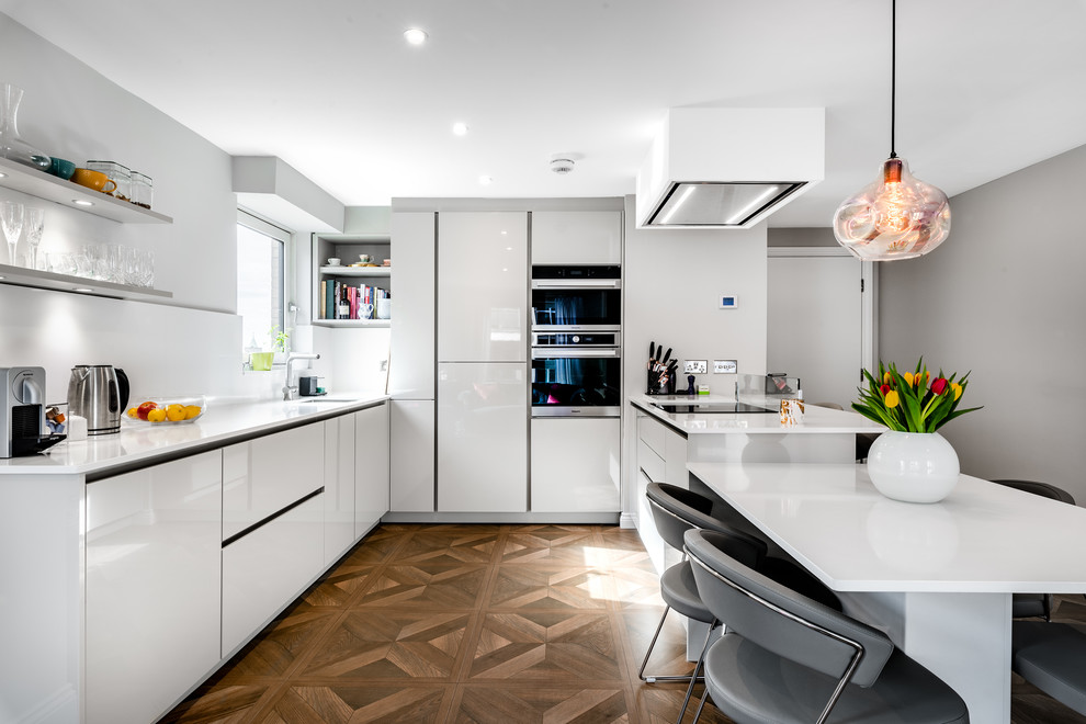 Contemporary u-shaped kitchen in London with an undermount sink, flat-panel cabinets, white cabinets, stainless steel appliances, medium hardwood floors, a peninsula, brown floor and white benchtop.