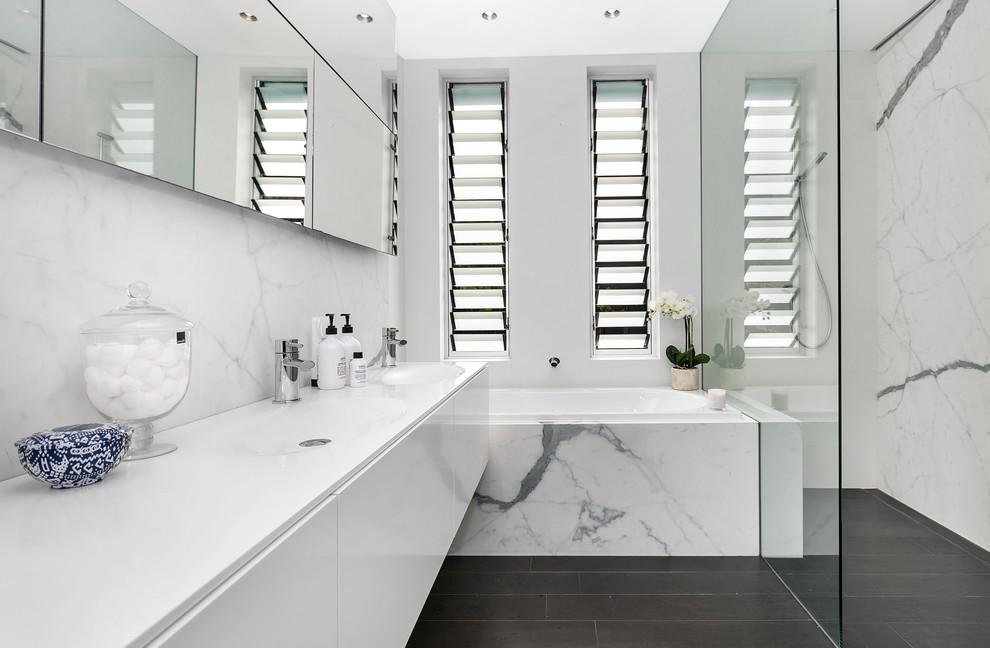 This is an example of a mid-sized contemporary master bathroom in Sydney with flat-panel cabinets, white cabinets, an alcove tub, an alcove shower, white tile, marble, white walls, a wall-mount sink, limestone benchtops, black floor and an open shower.