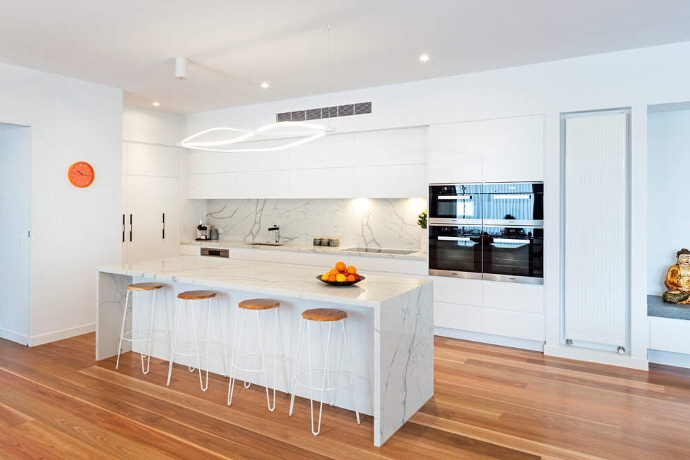 This is an example of a contemporary kitchen in Melbourne with an undermount sink, flat-panel cabinets, white cabinets, white splashback, stainless steel appliances, medium hardwood floors, with island, brown floor and white benchtop.