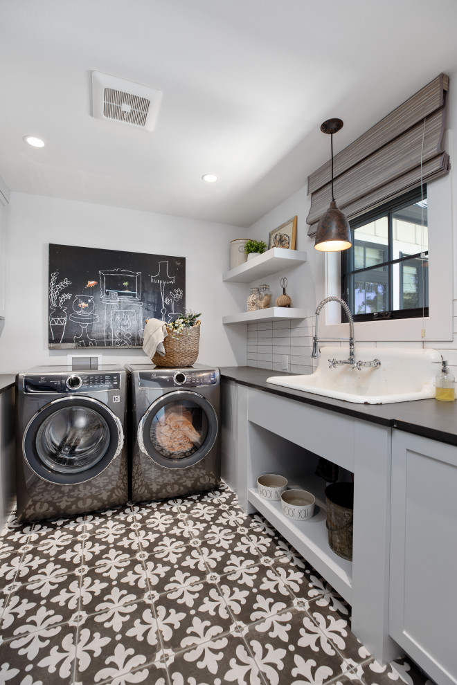 Inspiration for a mid-sized country single-wall dedicated laundry room in Portland with a farmhouse sink, shaker cabinets, grey cabinets, quartz benchtops, white splashback, ceramic splashback, white walls, ceramic floors, a side-by-side washer and dryer, black floor and black benchtop.