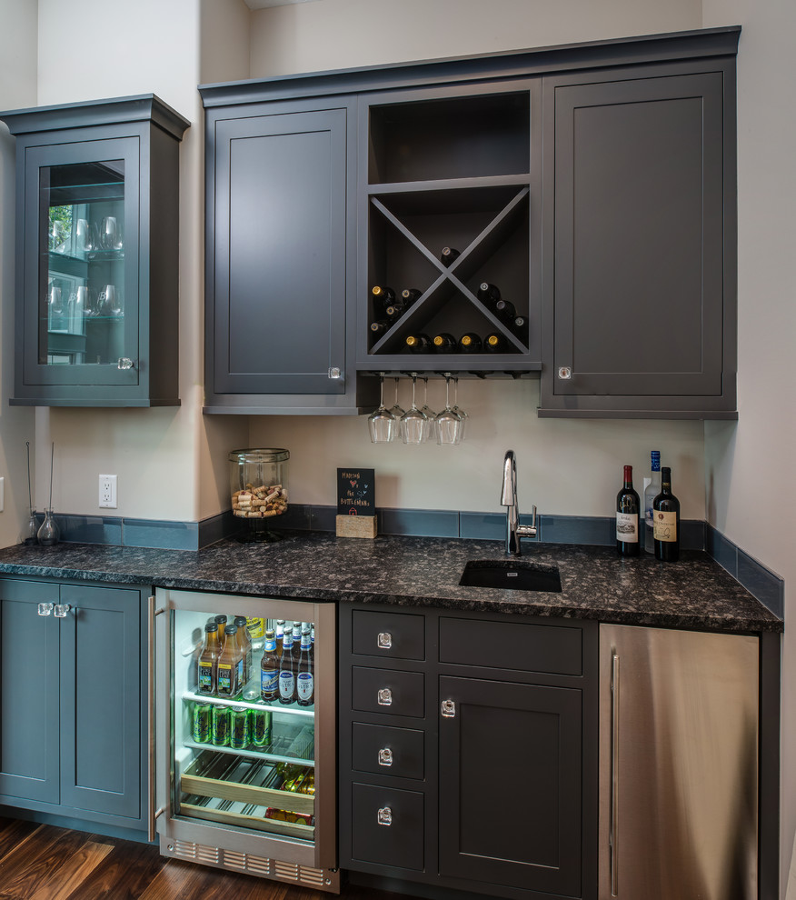 Example of a small transitional single-wall medium tone wood floor wet bar design with an undermount sink, shaker cabinets, gray cabinets, granite countertops, gray backsplash and glass tile backsplash