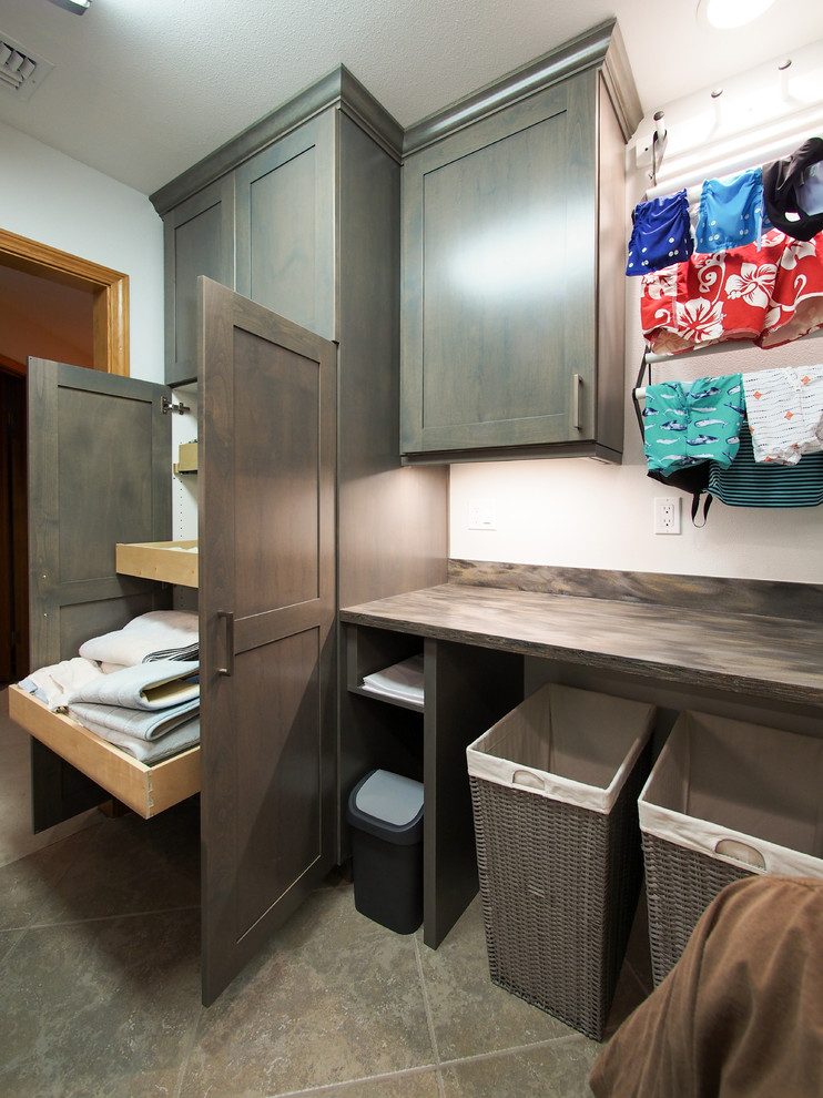 Mid-sized transitional galley dedicated laundry room in Seattle with an undermount sink, recessed-panel cabinets, dark wood cabinets, solid surface benchtops, blue walls, porcelain floors, a side-by-side washer and dryer, brown splashback, beige floor and brown benchtop.