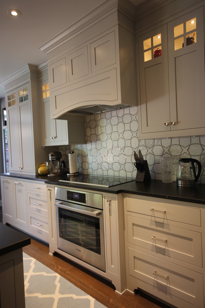 Photo of a mid-sized contemporary l-shaped eat-in kitchen in Montreal with a farmhouse sink, beaded inset cabinets, grey cabinets, solid surface benchtops, grey splashback, panelled appliances, dark hardwood floors, with island, brown floor, black benchtop and marble splashback.