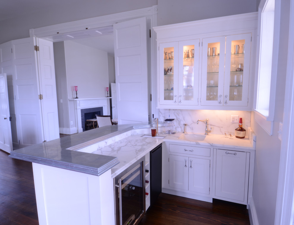 This is an example of a mid-sized transitional l-shaped wet bar in Baltimore with an undermount sink, shaker cabinets, white cabinets, marble benchtops, white splashback, stone slab splashback, dark hardwood floors and brown floor.