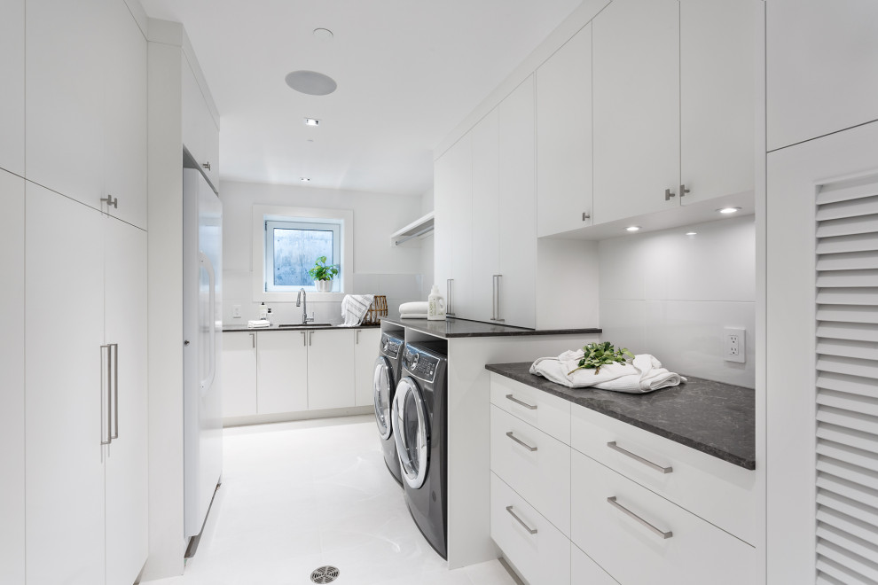 This is an example of an expansive contemporary galley dedicated laundry room in Vancouver with a drop-in sink, flat-panel cabinets, white cabinets, quartzite benchtops, white walls, vinyl floors, a side-by-side washer and dryer, white floor and black benchtop.