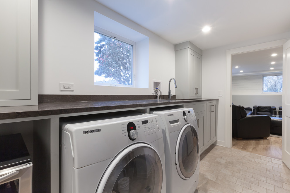 Mid-sized transitional single-wall utility room in Chicago with an undermount sink, flat-panel cabinets, grey cabinets, granite benchtops, grey walls, ceramic floors, a side-by-side washer and dryer, beige floor and black benchtop.