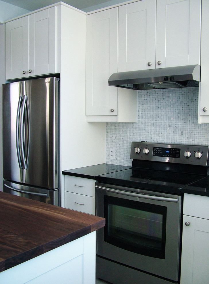 Photo of a mid-sized transitional l-shaped open plan kitchen in Montreal with shaker cabinets, white cabinets, grey splashback, stainless steel appliances, ceramic floors, an undermount sink, wood benchtops, mosaic tile splashback and with island.