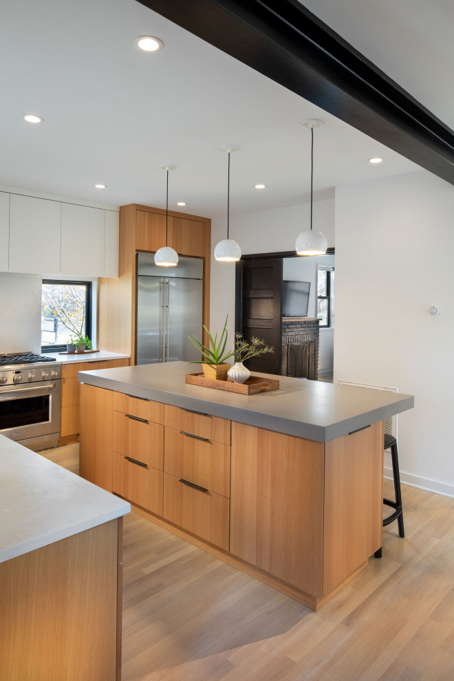 This is an example of a large contemporary l-shaped eat-in kitchen in DC Metro with an undermount sink, flat-panel cabinets, light wood cabinets, quartz benchtops, white splashback, stone slab splashback, stainless steel appliances, light hardwood floors, with island, brown floor and grey benchtop.