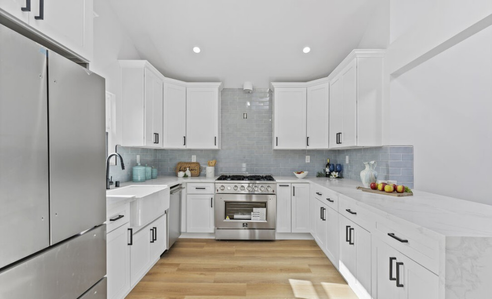 Photo of a contemporary u-shaped kitchen in Los Angeles with a farmhouse sink, shaker cabinets, white cabinets, quartz benchtops, grey splashback, ceramic splashback, stainless steel appliances, light hardwood floors, brown floor, white benchtop and vaulted.