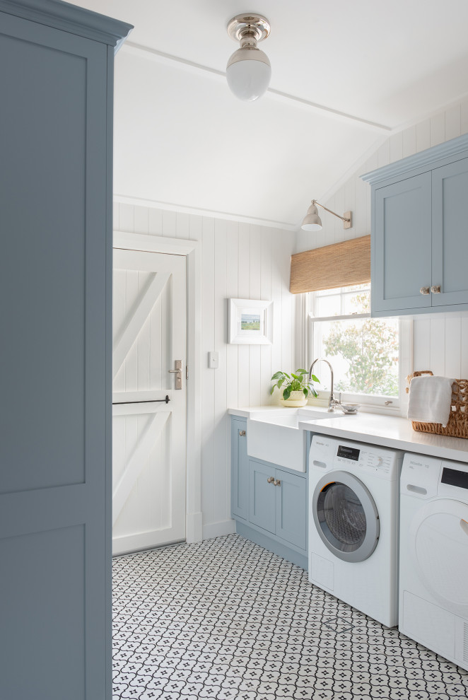 Design ideas for a mid-sized transitional galley utility room in Perth with a farmhouse sink, shaker cabinets, blue cabinets, a side-by-side washer and dryer, white benchtop, white walls, multi-coloured floor and planked wall panelling.