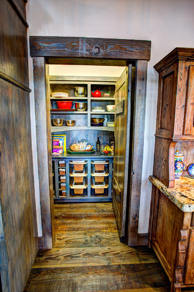Example of a large mountain style l-shaped dark wood floor eat-in kitchen design with a farmhouse sink, flat-panel cabinets, distressed cabinets, granite countertops, beige backsplash, stone tile backsplash, black appliances and an island