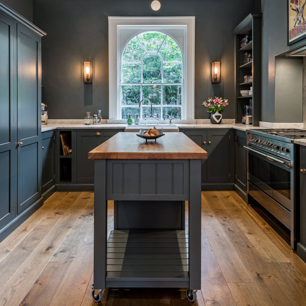 This is an example of a transitional u-shaped kitchen in London with a farmhouse sink, shaker cabinets, grey cabinets, stainless steel appliances, medium hardwood floors, with island, brown floor, white benchtop and grey splashback.
