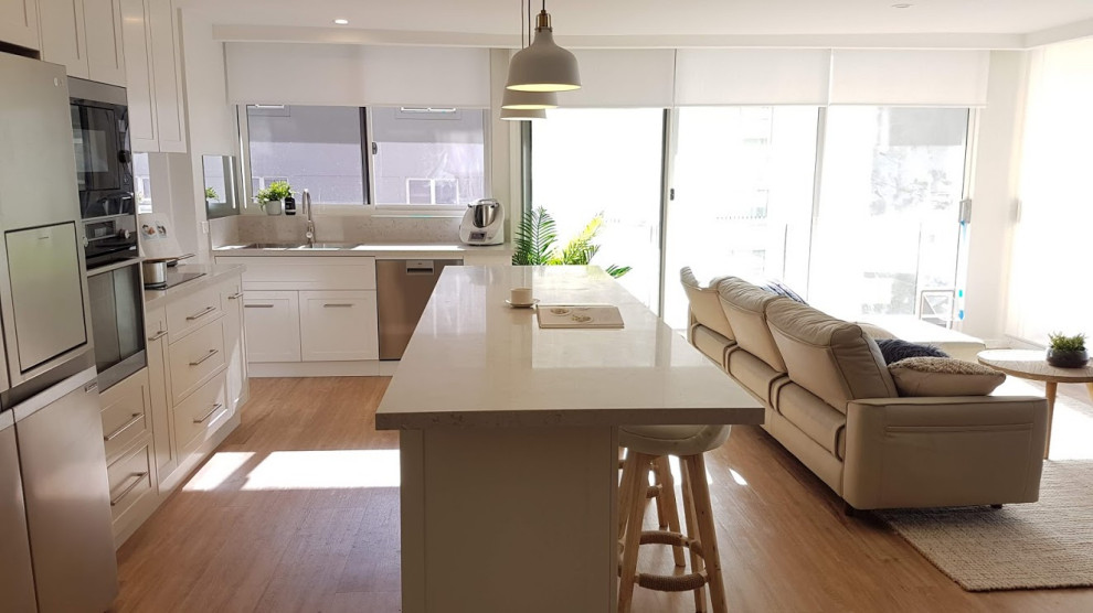 Small contemporary l-shaped open plan kitchen in Wollongong with a drop-in sink, shaker cabinets, white cabinets, quartz benchtops, metallic splashback, mirror splashback, stainless steel appliances, vinyl floors, with island, beige floor and white benchtop.