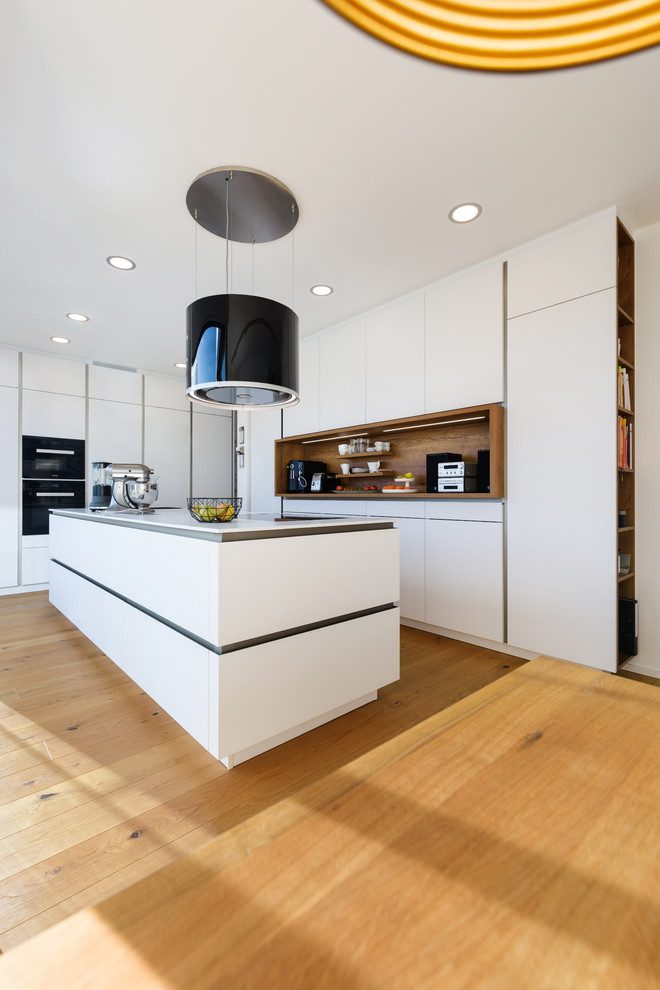 Design ideas for a mid-sized contemporary l-shaped open plan kitchen in Stuttgart with an integrated sink, flat-panel cabinets, white cabinets, solid surface benchtops, white splashback, timber splashback, black appliances, painted wood floors, with island and brown floor.