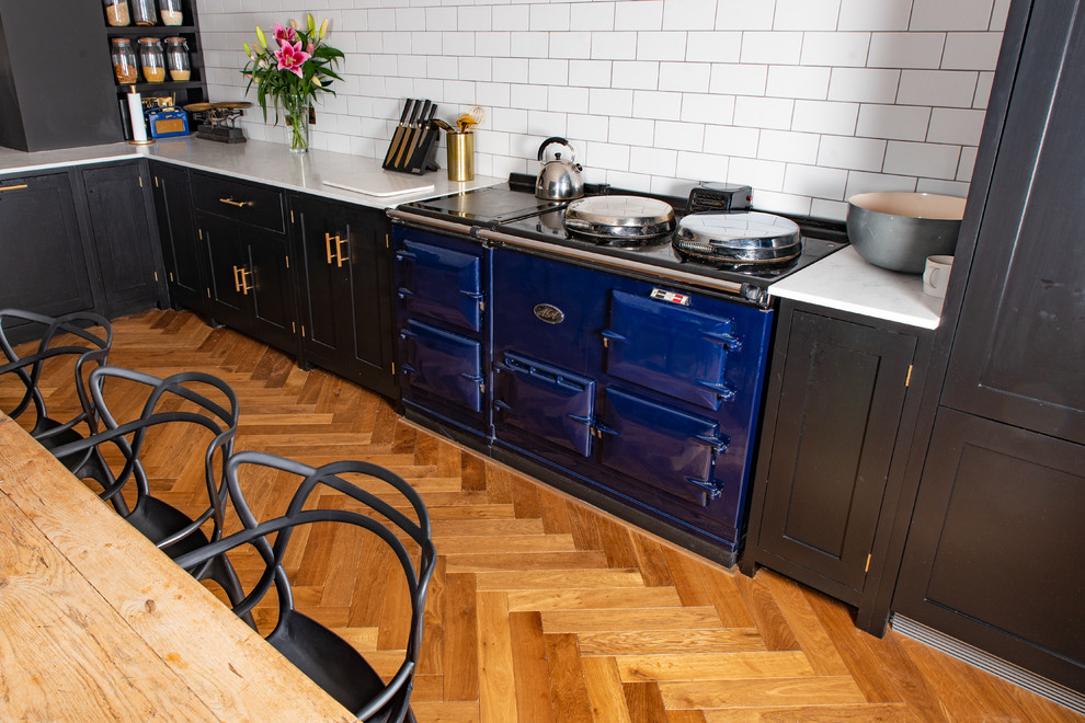 Photo of a rustic l-shaped kitchen/diner in Cambridgeshire with medium hardwood flooring, a belfast sink, recessed-panel cabinets, black cabinets, marble worktops, white splashback, porcelain splashback, black appliances, a breakfast bar, brown floors and white worktops.