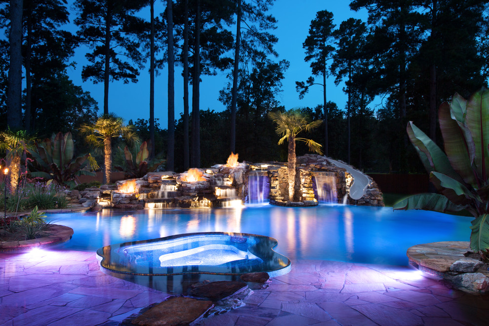 Photo of an expansive tropical backyard custom-shaped natural pool in New Orleans with natural stone pavers and a hot tub.