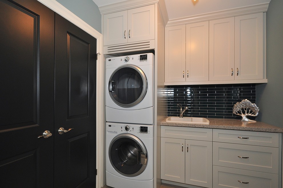 Design ideas for a mid-sized transitional single-wall dedicated laundry room in Toronto with a drop-in sink, shaker cabinets, white cabinets, granite benchtops, grey walls, ceramic floors, a stacked washer and dryer and beige floor.