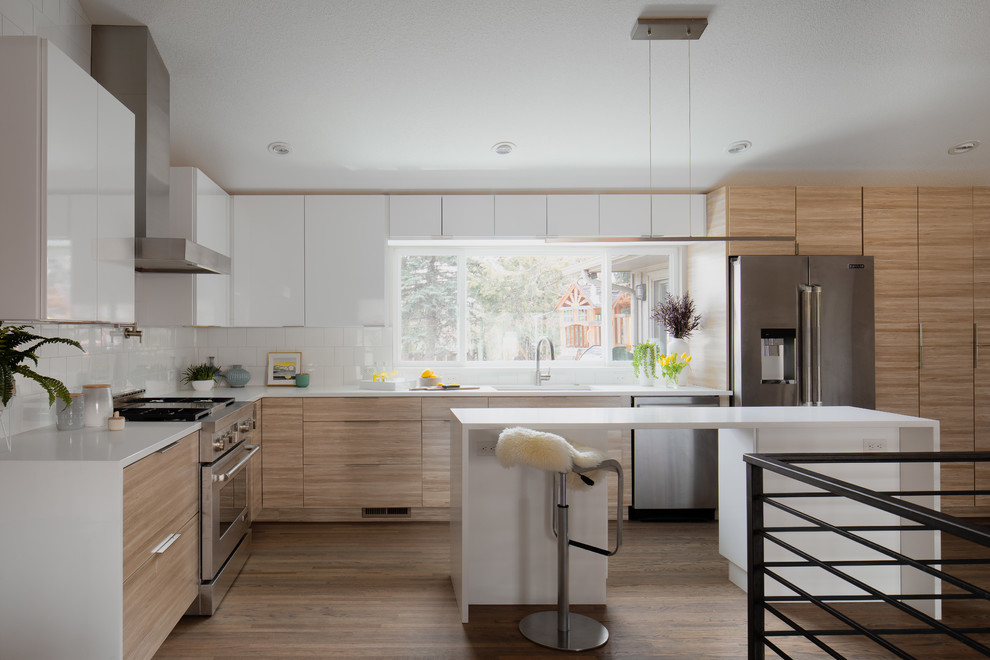 Mid-sized contemporary l-shaped kitchen in Denver with an undermount sink, flat-panel cabinets, light wood cabinets, window splashback, stainless steel appliances, light hardwood floors, with island and white benchtop.