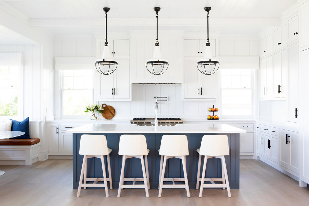 Large beach style u-shaped eat-in kitchen in New York with shaker cabinets, white cabinets, white splashback, stainless steel appliances, light hardwood floors, with island, beige floor, white benchtop, a farmhouse sink, quartz benchtops and timber splashback.