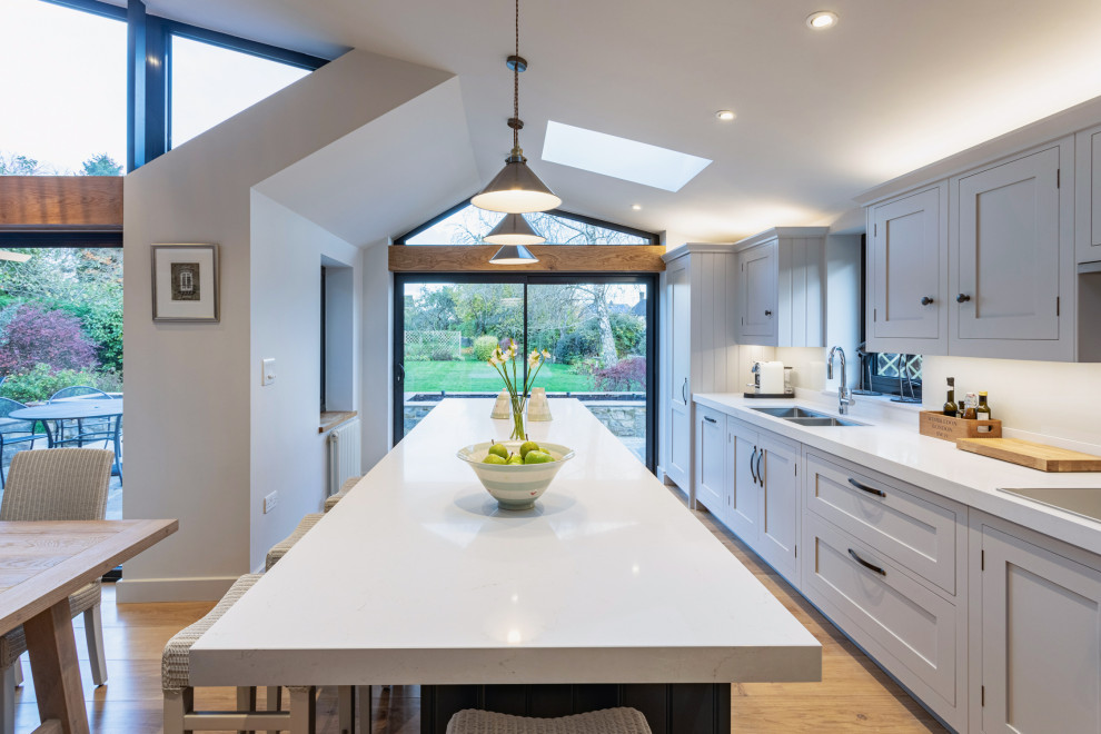 Inspiration for a transitional single-wall open plan kitchen in Gloucestershire with shaker cabinets, with island, an undermount sink, grey cabinets, panelled appliances, medium hardwood floors, brown floor and white benchtop.