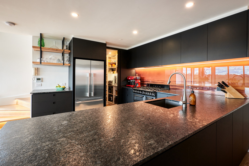 Пример оригинального дизайна: п-образная кухня-гостиная среднего размера в стиле модернизм с плоскими фасадами, черными фасадами, столешницей из кварцевого агломерата, оранжевым фартуком, фартуком из стекла, черной техникой и черной столешницей