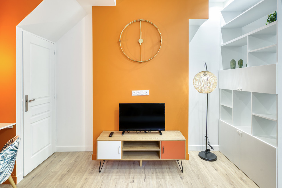 Пример оригинального дизайна: маленькая открытая гостиная комната в стиле лофт с с книжными шкафами и полками, оранжевыми стенами, светлым паркетным полом и отдельно стоящим телевизором для на участке и в саду