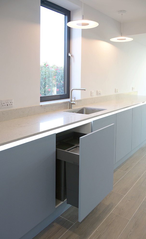 This is an example of a mid-sized contemporary open plan kitchen in Cornwall with flat-panel cabinets, blue cabinets, quartz benchtops, grey splashback, glass sheet splashback, stainless steel appliances and grey benchtop.