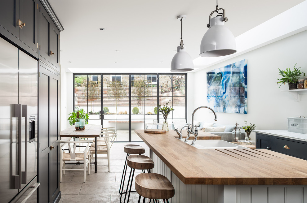 Inspiration for a large scandinavian galley eat-in kitchen in London with a farmhouse sink, recessed-panel cabinets, wood benchtops, stainless steel appliances, with island, black cabinets, white splashback and grey floor.