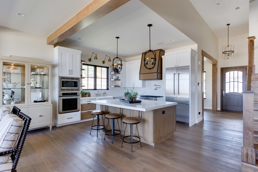 Inspiration for a country l-shaped kitchen in Salt Lake City with a farmhouse sink, shaker cabinets, white cabinets, quartzite benchtops, white splashback, subway tile splashback, stainless steel appliances, light hardwood floors, with island and white benchtop.