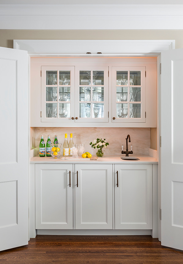 Photo of a small traditional single-wall wet bar in Minneapolis with an undermount sink, glass-front cabinets, white cabinets, marble benchtops, white splashback, stone slab splashback and medium hardwood floors.