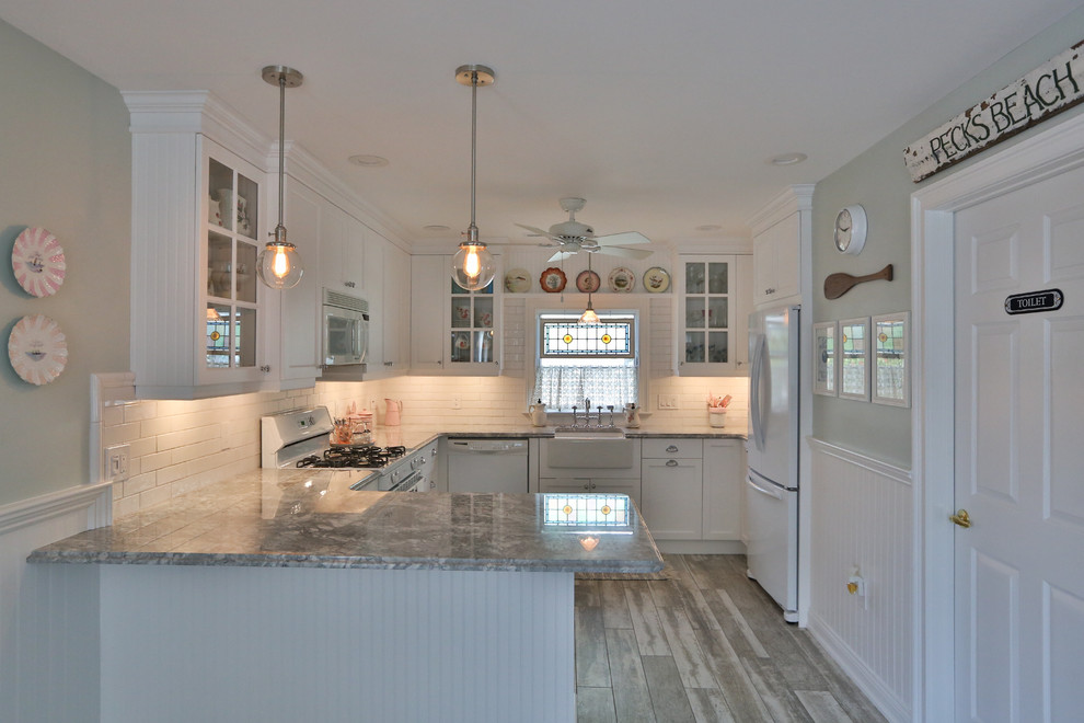 This is an example of a small beach style l-shaped kitchen in Philadelphia with a farmhouse sink, shaker cabinets, white cabinets, marble benchtops, white splashback, subway tile splashback, white appliances, ceramic floors and with island.