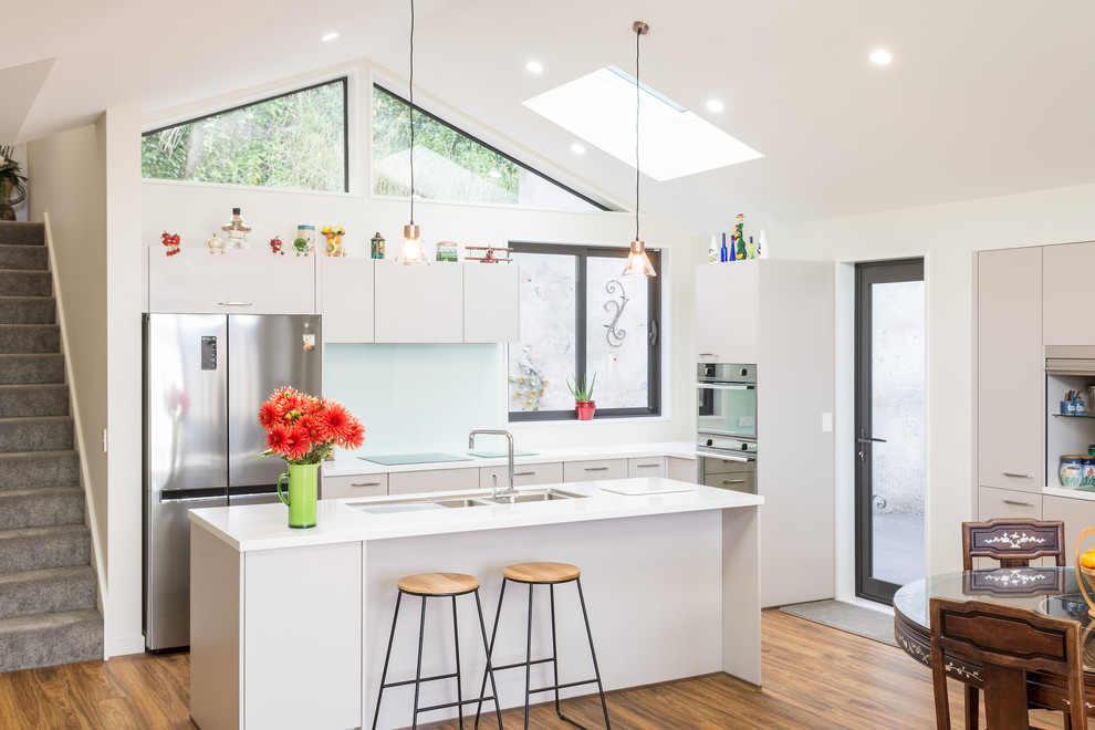 Contemporary l-shaped open plan kitchen in Christchurch with a double-bowl sink, flat-panel cabinets, white cabinets, glass sheet splashback, stainless steel appliances, dark hardwood floors, with island and white benchtop.