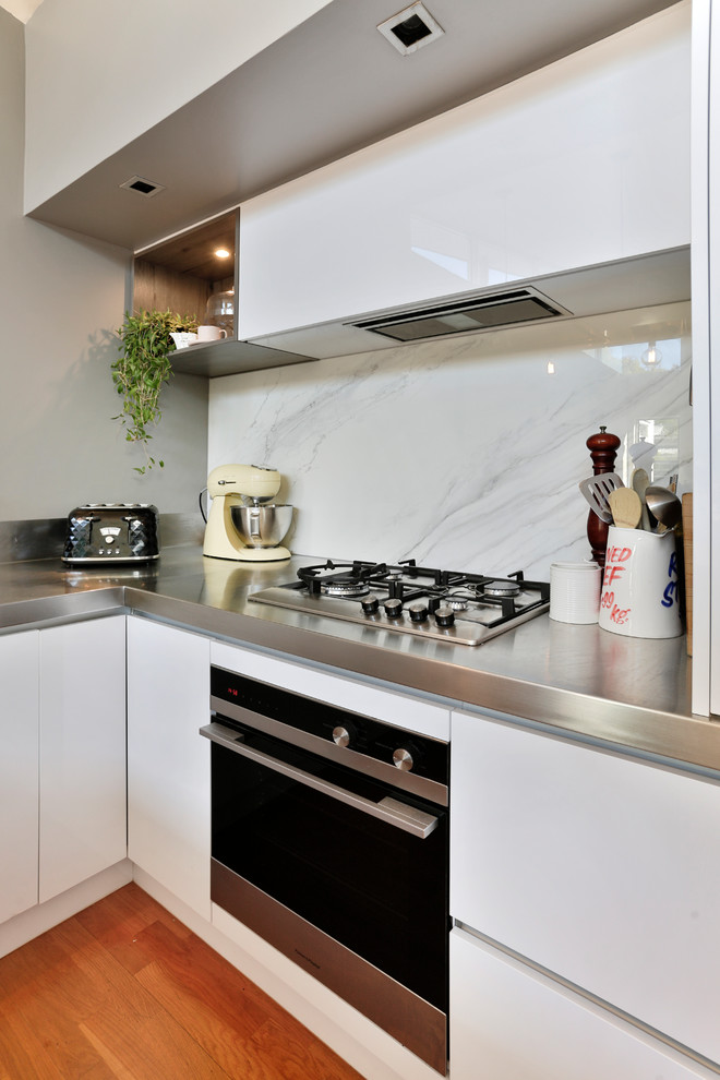 Inspiration for a large industrial u-shaped open plan kitchen in Auckland with a double-bowl sink, flat-panel cabinets, white cabinets, stainless steel benchtops, white splashback, glass sheet splashback, stainless steel appliances, medium hardwood floors, with island and multi-coloured floor.